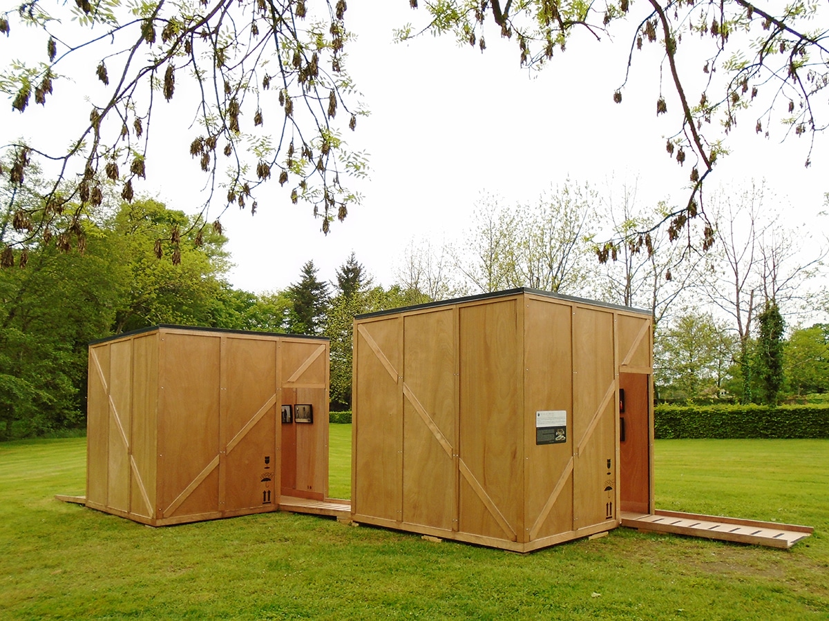 Vue extérieure de 2 modules d’expo, en bois, rappelant des caisses de transport, posés dans le parc d’une abbaye