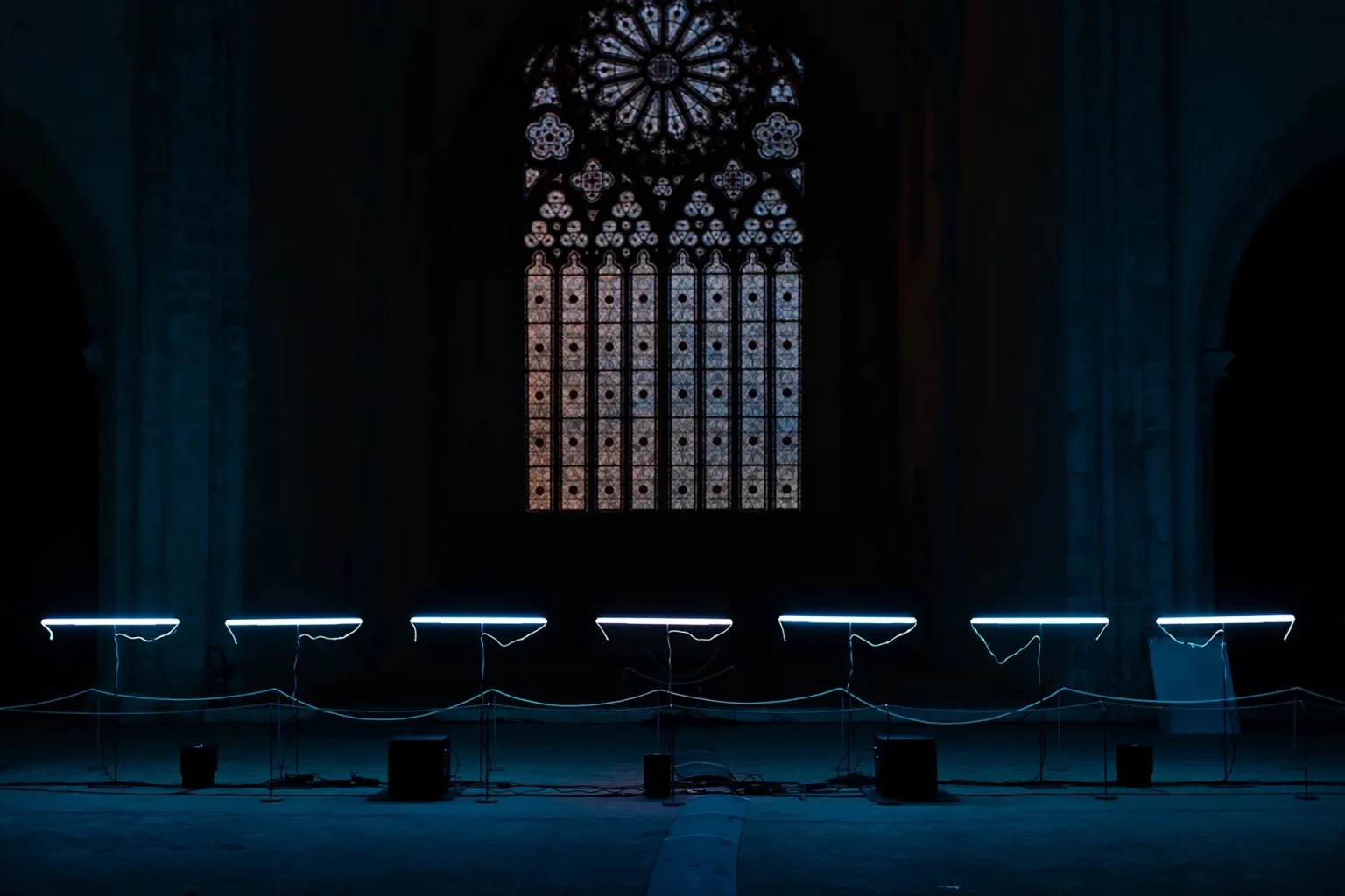 Échos : ligne de tubes lumineux dans la nef sombre d'une église, rosace en fond